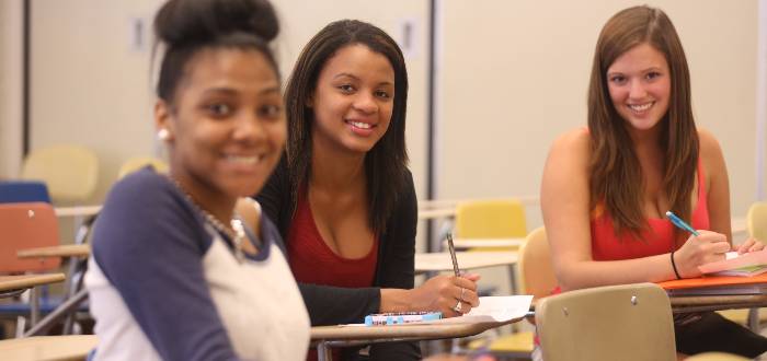 Three smiling students sit in a classroom and look at the camera.